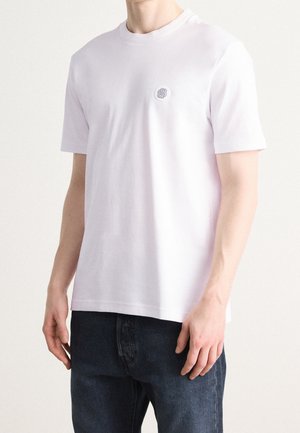 Biała krótkorękawowa koszulka z bawełny z okrągłym dekoltem, ozdobiona małym okrągłym logo na piersi. W komplecie ciemne dżinsy.