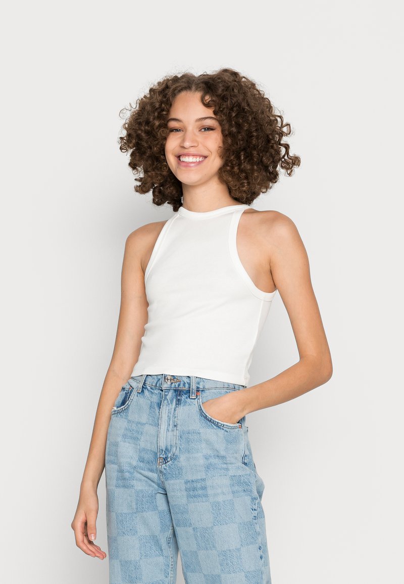 Top cropped branco sem mangas, de design ajustado com um decote alto. Usado com jeans de cintura alta xadrez azul claro. Fundo plano.