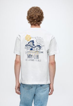 Persoană care poartă un tricou alb cu un grafic albastru și galben ce reprezintă valuri, soare, înotător și textul “Swim Club La Grand Plage” pe spate.