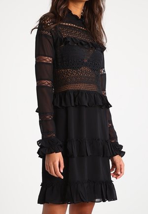 Kobieta ubrana w czarną sukienkę z długim rękawem z koronkowymi panelami, falbanami na rękawach, piersiach, tali i na dole, na białym tle.