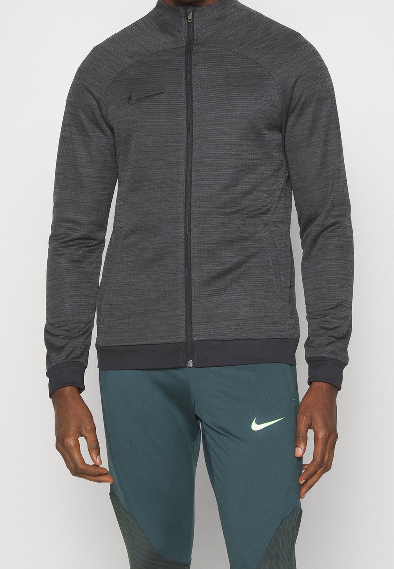 Šedá sportovní bunda na zip s texturovanou tkaninou, přiléhavým střihem a pružnými manžetami. Obsahuje černé logo Nike na hrudi.