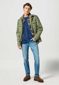 Chaqueta militar verde oliva, camiseta de cuello redondo azul marino, camisa a cuadros por debajo, jeans de mezclilla azul claro y zapatos informales marrones.
