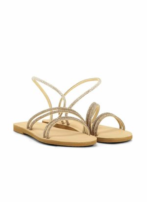 Sandales plates beige avec des fines lanières dorées étincelantes qui croisent sur les orteils et autour de la cheville sur fond blanc.