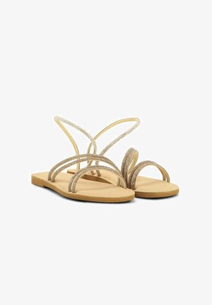 Sandales plates beige avec des fines lanières dorées étincelantes qui croisent sur les orteils et autour de la cheville sur fond blanc.