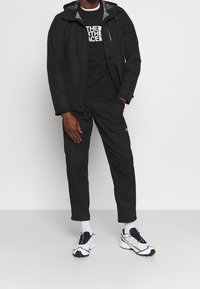 Svart vattentät jacka med huva, i kombination med en svart sweatshirt och smalpassande svarta byxor. Vita och svarta sneakers fulländar outfiten.