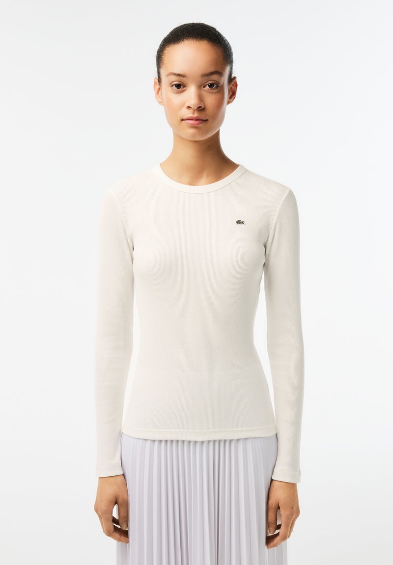 White long-sleeve top with a ribbed texture and round neckline, featuring a small green logo on the left side. Smooth fabric finish.
