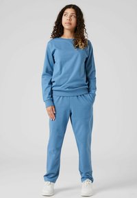 Giovane con capelli ricci che indossa una felpa blu e pantaloni abbinati, in piedi con le mani in tasca, che guarda in alto a destra.