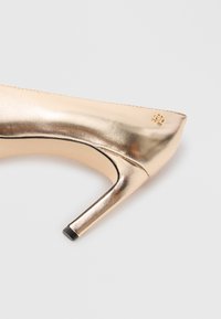 Metallic guld högklackad sko med spetsig tå, slät yta och en svart detalj vid klacken. Har en liten logotyp nära klacken.