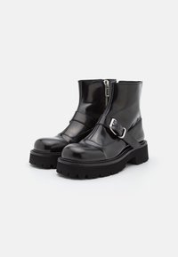 Botines de charol negro con cremallera lateral, puntera afilada, suela de caucho gruesa y una correa decorativa con hebilla plateada.