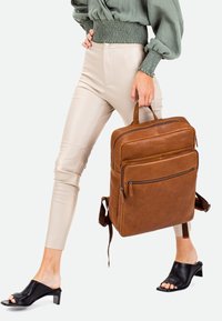 Mochila de cuero marrón con dos cremalleras frontales, asa superior y correas de hombro ajustables. Textura suave con un diseño minimalista.