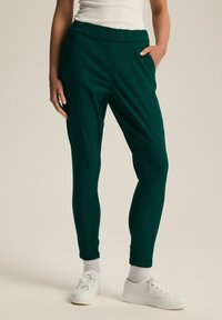 Zielone, elastyczne spodnie dresowe z gumką w talii, bocznymi kieszeniami i zwężanym krojem. W zestawie z białymi sneakerami i skarpetkami.