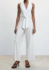 Mujer vestida con un mono sin mangas de rayas blancas con detalle de nudo delantero y pantalones de pierna ancha, combinado con sandalias de tacón marrones.