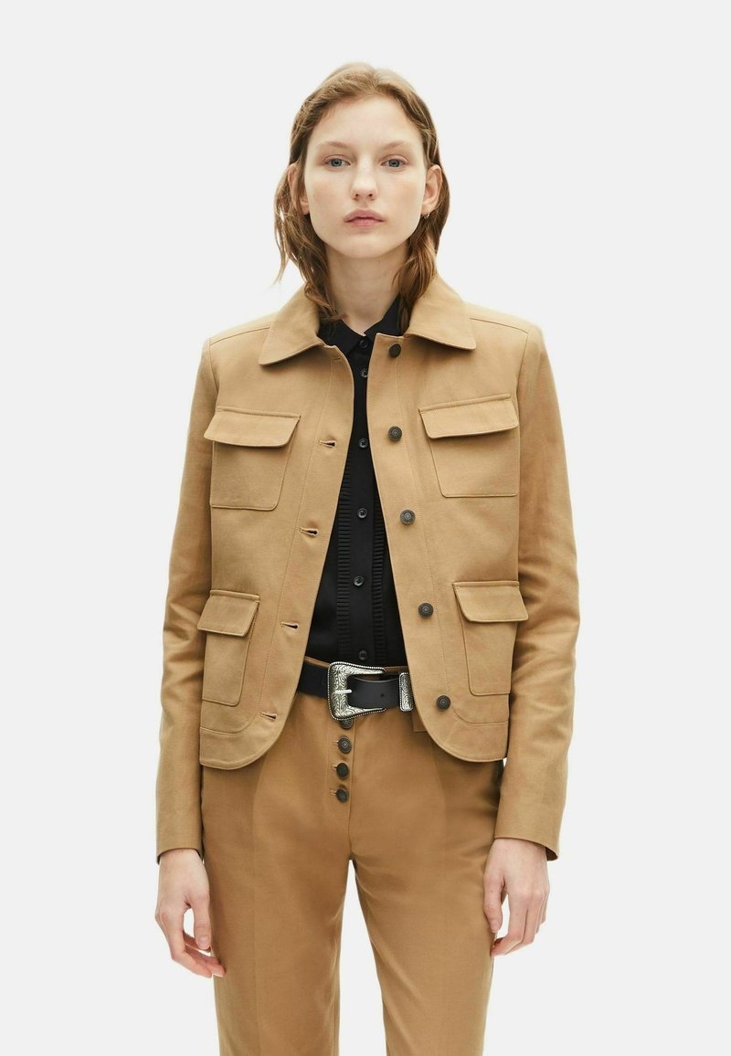 Veste en mélange de coton beige avec quatre poches à rabat, fermeture à bouton, col et design légèrement ajusté, portée sur une chemise noire et un pantalon.