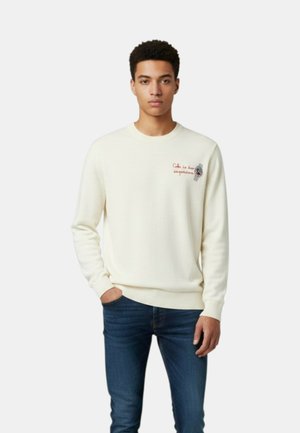 Giovane uomo con una mano in tasca che indossa un maglione bianco con un piccolo testo rosso e jeans blu scuro, in piedi davanti a uno sfondo semplice.