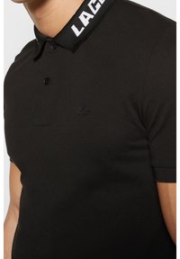Polo noir à manches courtes avec logo crocodile brodé sur la poitrine et texte de marque "LACOSTE" en blanc sur le col.