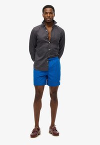 Camicia nera con colletto, pantaloni corti blu, calzature marroni. Modello in piedi con le mani dietro la schiena, espressione neutra, sfondo semplice.