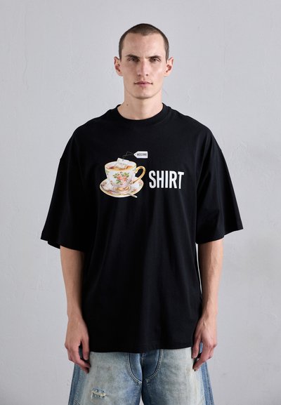 Sort oversized t-shirt med et floral te-kop og underkop grafik, med ordet "SHIRT" trykt i hvid. Fremstillet af bomuld, afslappet pasform.