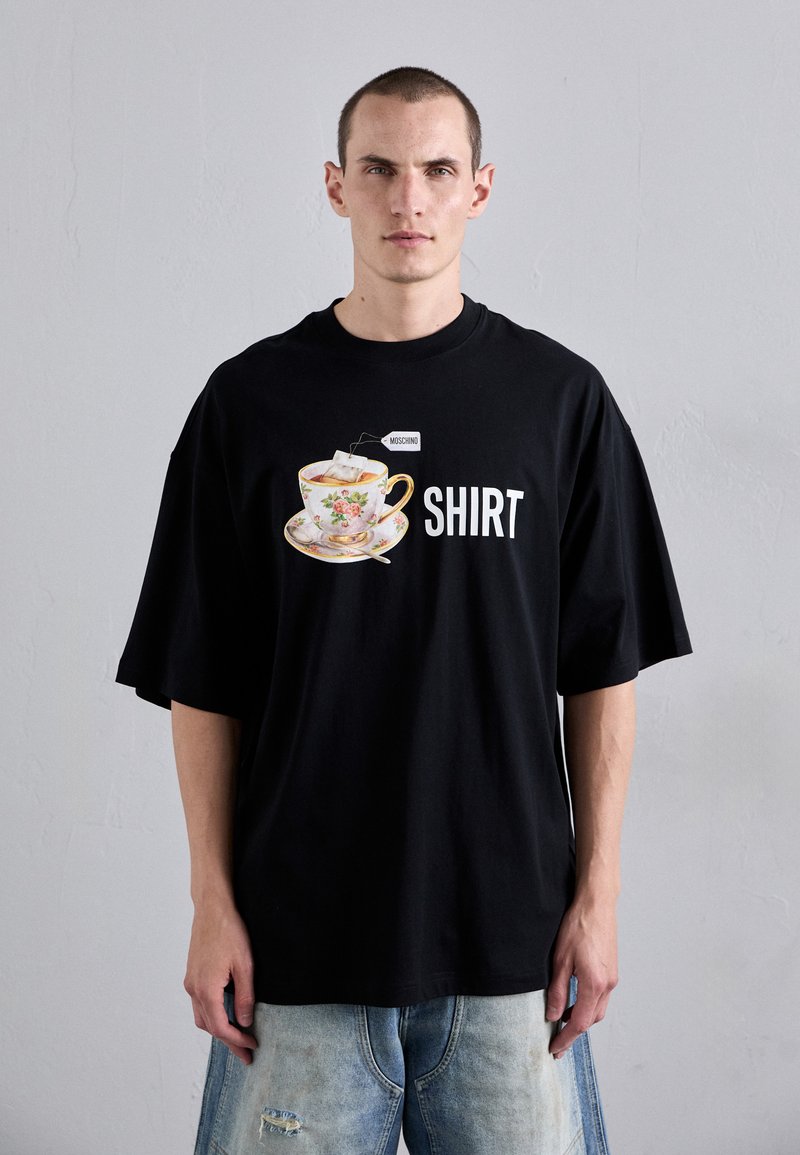 T-shirt noir oversize avec un graphique de tasse à thé florale et sa soucoupe, sur lequel le mot "SHIRT" est imprimé en blanc. Fabriqué en coton, coupe décontractée.