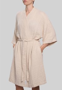 Šviesiai smėlio spalvos, tekstūruotas chalatas su apvyniojamu dizainu, plačiomis rankovėmis ir diržu. Yra šoninės kišenės ir etiketė hemelyje.