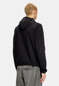 Veste à capuche noire en tissu léger et texturé ; dotée de poignets et d'un ourlet élastiques, accompagnée de pantalons gris ajustés.