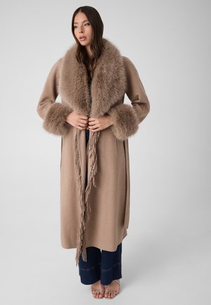 Dans un studio, le mannequin porte un long manteau taupe avec un col et des poignets en fourrure moelleuse, des liens à franges, un jean large et des sandales à brides.