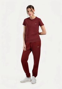 Bordowy zestaw sportowy składający się z krótkiego t-shirtu i zwężanych spodni. Tkanina wydaje się gładka z delikatnym połyskiem; zawiera detal z logo.