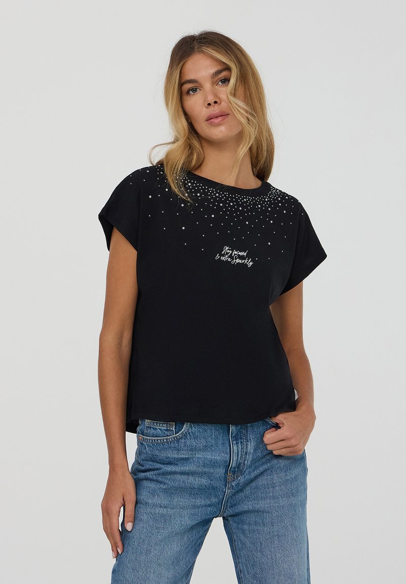 Maglietta nera a maniche corte con scollo rotondo, decorata con borchie argentate e una stampa di testo bianca, abbinata a jeans azzurri chiari.
