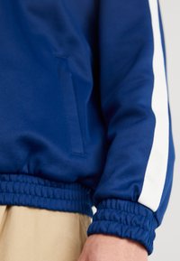 Henkilö, joka on pukeutunut siniseen takkiin, jossa on valkoinen raita hihassa, ja vaaleisiin housuihin, joissa näkyvät takin joustavat hihansuut ja alahelma.