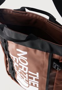 Καφέ και μαύρη τσάντα tote The North Face με φαρδιές λαβές και ανοιχτό πάνω μέρος που δείχνει την εσωτερική επένδυση και την τσέπη με φερμουάρ.