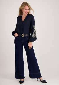 Marineblauw overhemd met knopen en geborduurde mouwen, gecombineerd met wijde donkerblauwe spijkerbroek en een zwarte riem. Zwarte schoenen met hak en strikdetail.