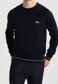 Muž v upnutém černém roláku s bílým logem "BOSS Hugo Boss" na hrudi a s tmavými džíny, s rukama částečně v kapsách.