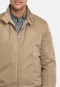 Chaqueta aislante color beige con cierre frontal de cremallera, cuello doblado y bolsillos laterales. El interior presenta un patrón a cuadros en verde y blanco.