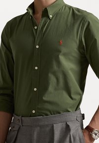 Groen overhemd met knopen en een op maat gemaakte kraag, voorzien van een klein rood logo. Gemaakt van gladde stof, gecombineerd met grijze broek en een horloge.