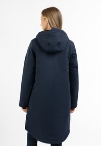Námořnický modrý parka kabát s kapucí, dlouhými rukávy a rovnými liniemi. Má hladký povrch a boční zipové kapsy.