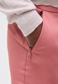 Χέρι με μανίκι ανοιχτό ροζ πουλόβερ ακουμπώντας μέσα σε φερμουάρ τσέπης ροζ φούστας ή παντελονιού σε λευκό φόντο.