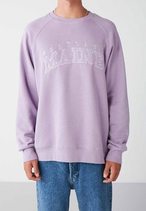 Person trägt einen lavendelfarbenen Pullover mit dem Schriftzug "Portland Maine" und blaue Jeans und steht vor einem schlichten hellen Hintergrund.