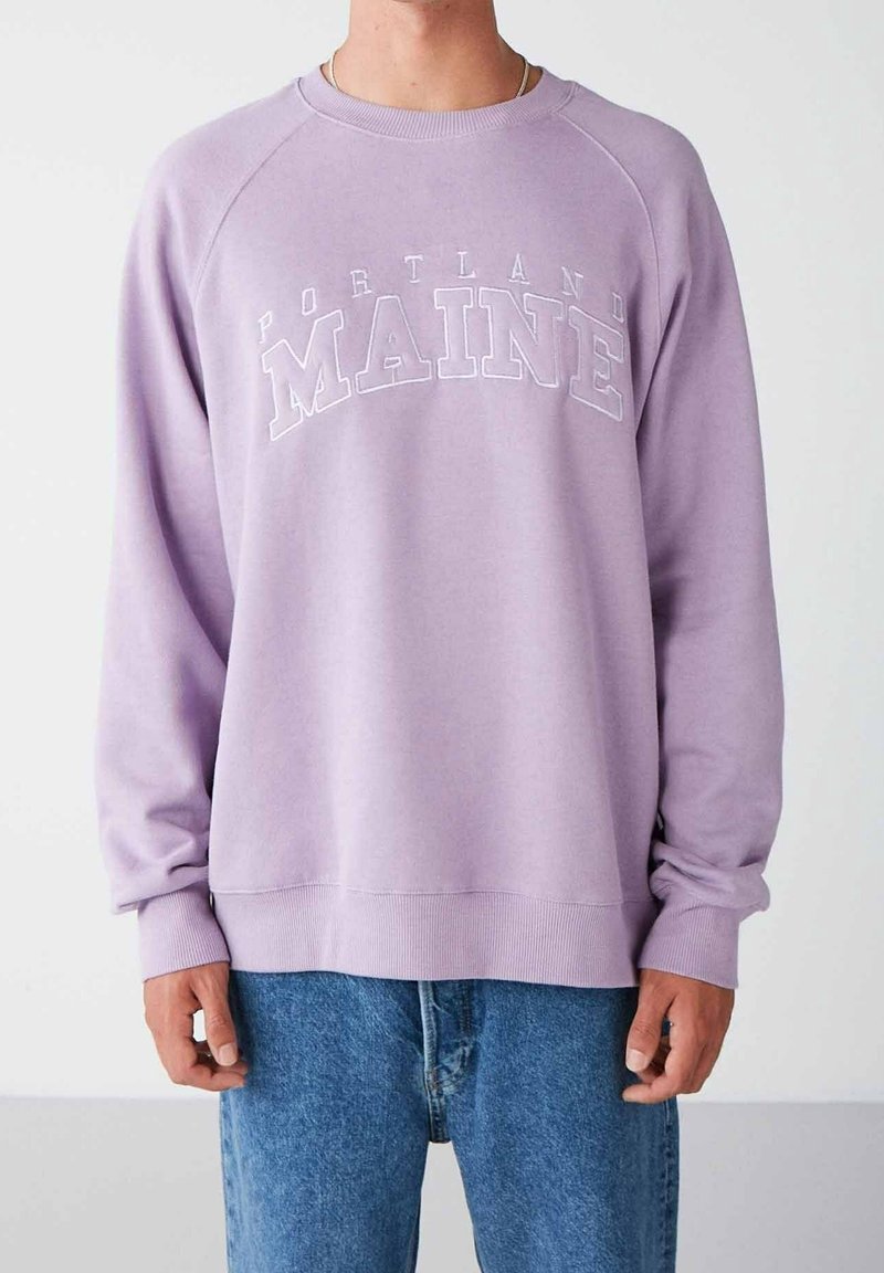 Personne portant un sweat lavande avec le texte « Portland Maine » et un jean bleu, debout devant un fond clair uni.