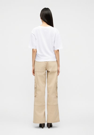 Γυναίκα με μακριά σκούρα μαλλιά, που φοράει ένα απλό άσπρο κοντομάνικο πουκάμισο και μπεζ παντελόνι cargo, στέκεται γυρισμένη με την πλάτη σε απλό φόντο.