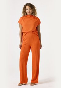 Oranje tweedelige outfit met een top met hoge hals en korte mouwen, en wijde pijpen met verticale naden. Glad materiaal en een ontspannen pasvorm.