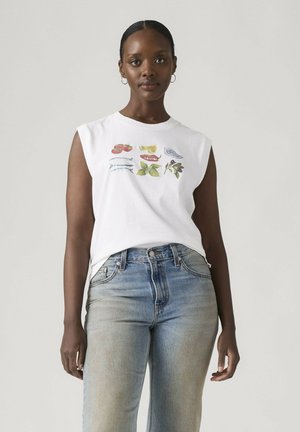 Joven mujer con camiseta blanca sin mangas con estampados de frutas y verduras y vaqueros azul claro, de pie frente a un fondo liso.