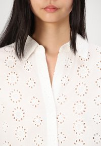 White shirt made of lightweight fabric, featuring embroidered floral patterns and eyelet cutouts. Classic collar and button closure detail.
