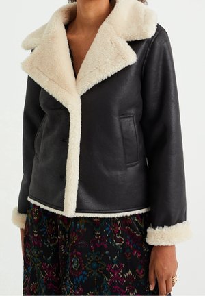 Czarna kurtka z sztucznej skóry z kremowym kołnierzem, mankietami i krawędzią obszytą futerkiem, noszona na wielobarwnej spódnicy z wzorem.