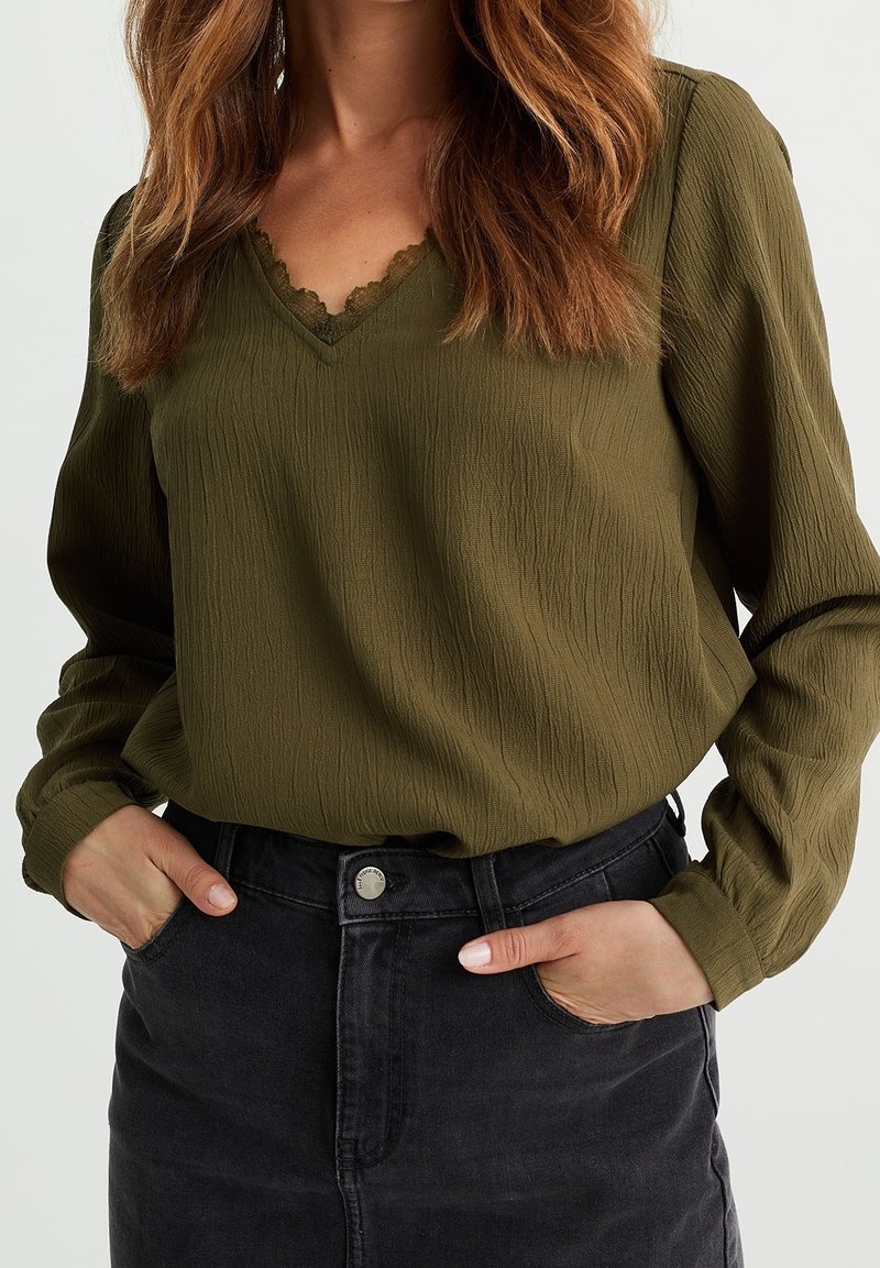 Olivgrön blus med texturerat tyg, V-ringning med spetsdetalj, kombinerad med en högmidjad svart jeanskjol med knapp.