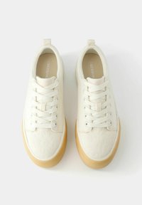 Białe trampki na sznurowadła z fakturowanej tkaniny i jasnobrązową gumową podeszwą, widziane z góry na jednolitym białym tle.