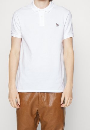 Person iført en enkel hvid kortærmet polo med et farverigt zebra-logo på brystet og brune læderbukser, der står mod en hvid baggrund.