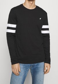 Svart långärmad T-shirt i bomull med vita ränder på ärmarna och en liten logotyp på bröstet. Bärs med blåjeans.