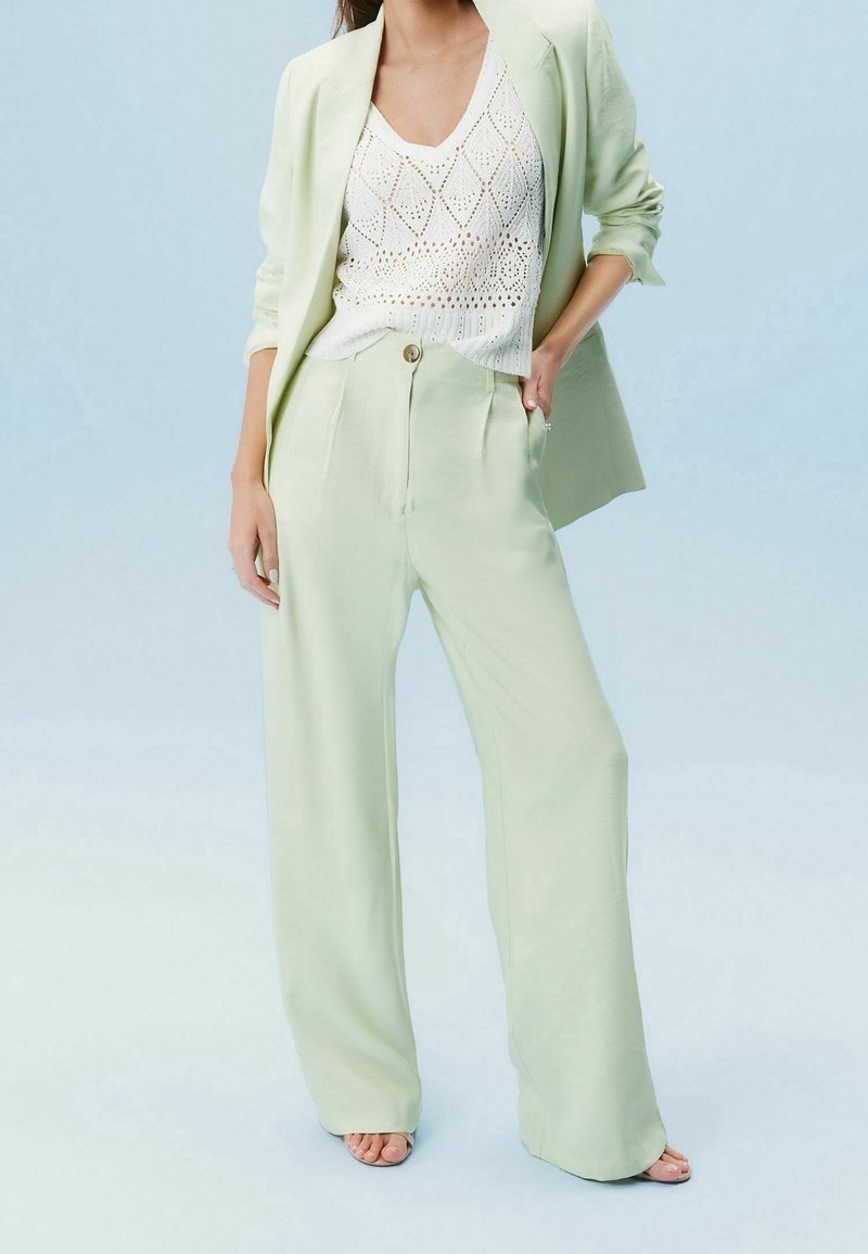 Femme portant un pantalon large vert clair et un blazer assorti avec un haut sans manches en maille blanche, debout avec une main dans la poche.