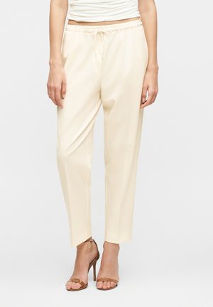 Donna che indossa pantaloni color crema a gamba stretta con vita elasticizzata e sandali marroni con tacco alto, in piedi davanti a uno sfondo semplice.