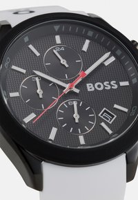 Black dial watch with three sub-dials, bold red second hand, white silicone strap, and black bezel. Features textured surface and date display.