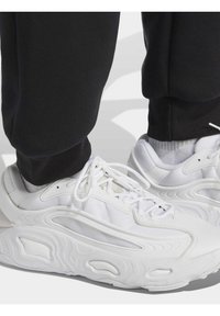 White athletic sneakers with a textured upper and smooth accents. Unique wavy sole design and white laces. Paired with black jogger-style pants.